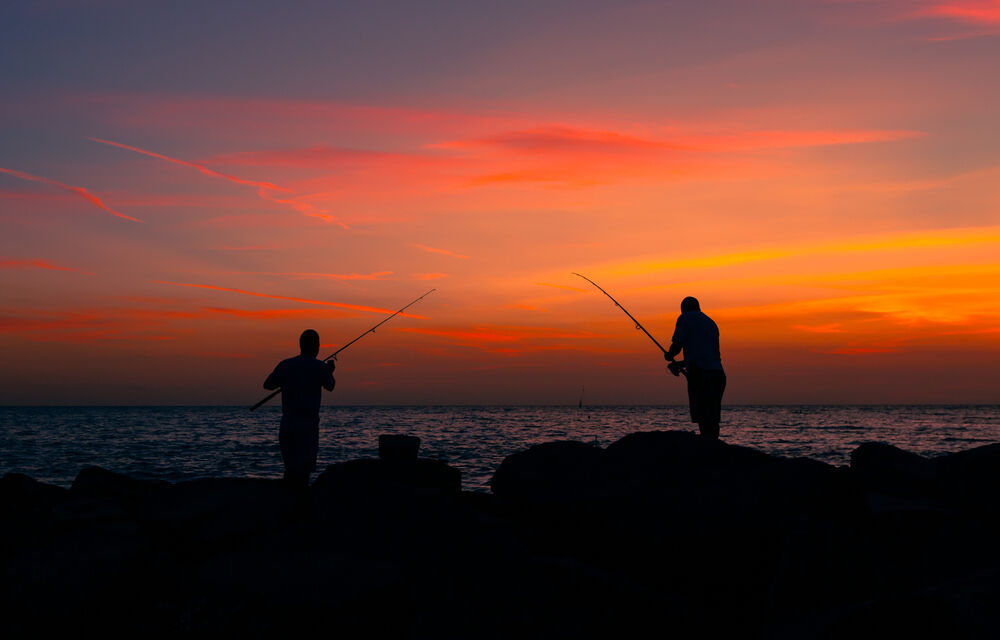 Two silhouetted fishermen stand on a rocky pier with a sunset over ocean water in the background.