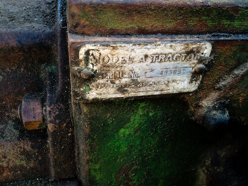 A portion of the body of a green tractor has grease and grime on it, along with a white maker plate bearing the words "Model A Tractor", a serial number, and a patent marking that reads "Patents Applied For".