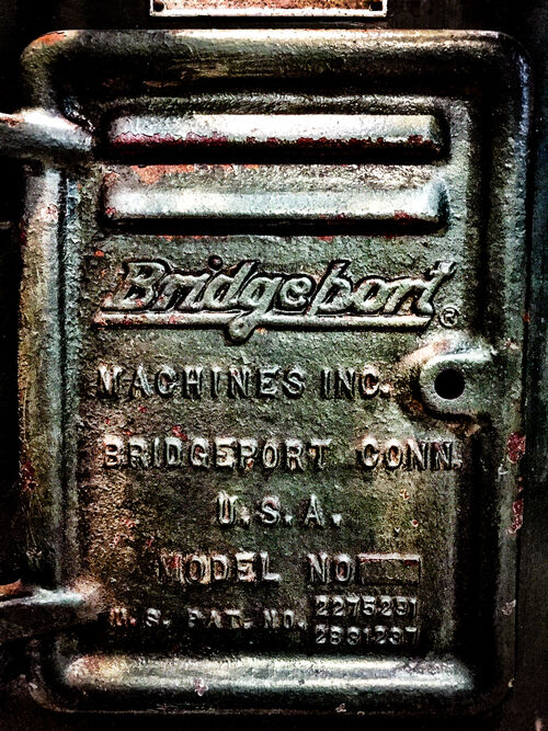 A metal column door from a Bridgeport milling machine fills the frame and includes the Bridgeport registered trademark, the words BRIDGEPORT CONN. U.S.A., a Model No. indicator, and a patent marking that lists to U.S. patent numbers.