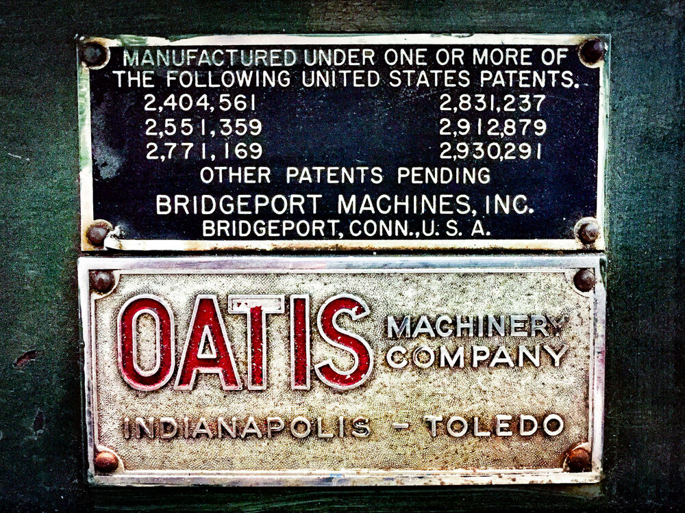 Two metal plates riveted to a machine are stacked vertically and fill the frame. The top plate is black with light lteering and lists a series of United States patent numbers, along with the text "Other Patents Pending" and "Bridgeport Machines, Inc.". The lower plate is yellowish-white in color and has raised lettering that reads "OATIS MACHINERY COMPANY INDIANAPOLIS - TOLEDO". The word "OATIS" is in red lettering, while all other lettering is the same color as the plate.