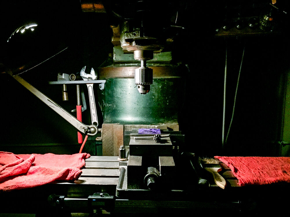 A workspace at a milling machine in includes shop towels and hand tools on the working surfacce of the machine. The room is dark and the milling machine is lit by a worklght attached to the column of the machine.