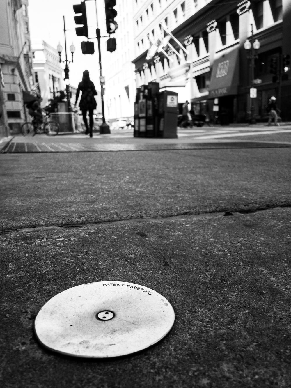 A black and white image shows a street scene with a cover of a sidewalk bait trap in the foreground with a marking for U. S. Patent No. 5,927,000. A woman walks away in the background.